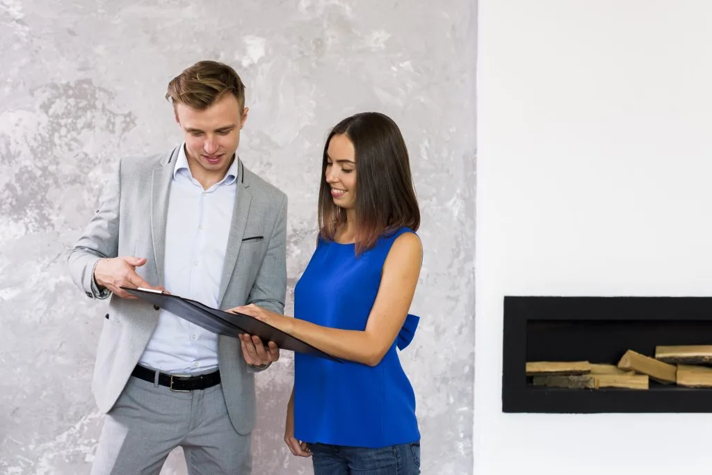 Un agente inmobiliario profesional con un traje gris hablando de los detalles de la propiedad con una clienta con una blusa azul dentro de una casa moderna.