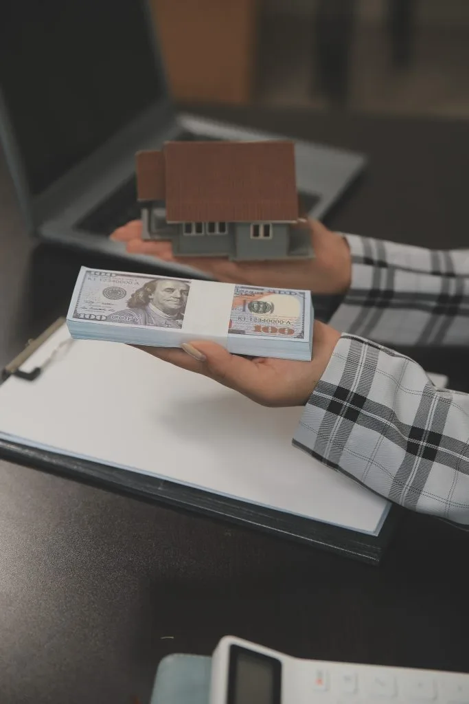 Real estate transaction concept with a person holding a house model and a stack of cash, symbolizing property investment and sales."