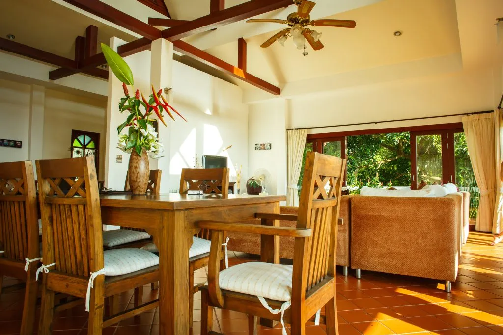 Bright, open-concept dining and living space featuring a wooden table, tropical floral arrangement, and large windows overlooking lush greenery in Phuket.