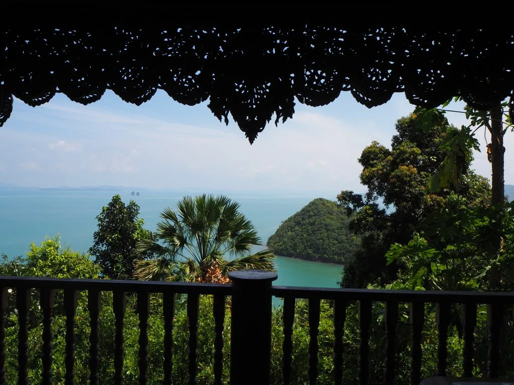 Scenic view of a turquoise sea and lush greenery seen through an ornate wooden balcony in Phuket.